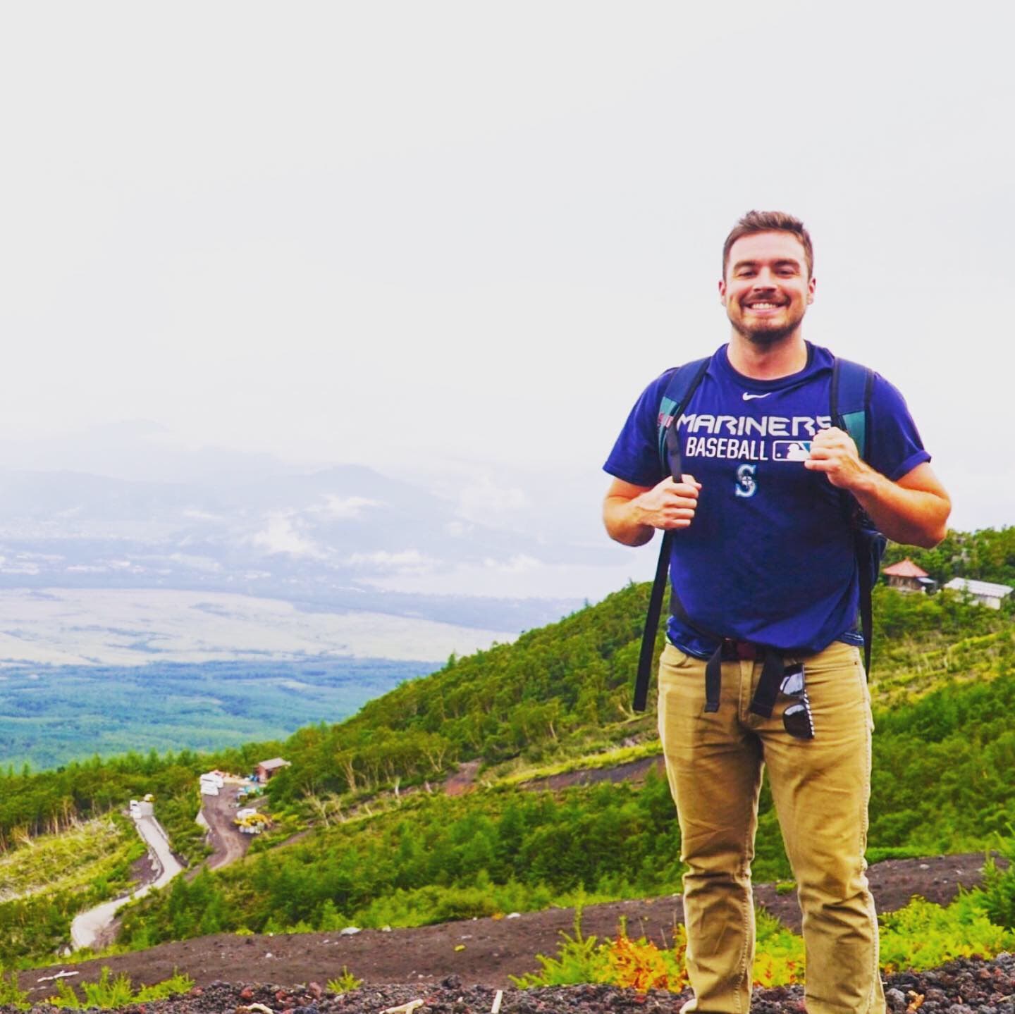 Bryce Henthorn at altitude on a hike, smiling toward the camera with a green valley and clouds behind